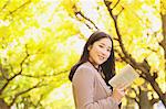Japanese woman with long hair looking at camera while holding a book with yellow leaves in the background