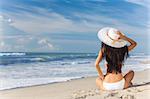 Ein sexy junge Brünette Frau oder Mädchen trägt einen weißen Bikini und Sonne Hut sitzt auf einem einsamen tropischen Strand mit einem blauen Himmel