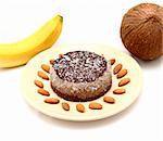 Home made cake on a brown plate covered with chocolate cream and coconut flakes on a white table with banana, almond seeds and coconut