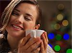 Young woman enjoying cup of hot beverage in front of Christmas lights