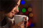 Happy young woman enjoying cup of hot beverage in front of Christmas lights