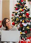 Thoughtful young woman with laptop sitting near Christmas tree