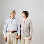 Senior adult couple holding hands and smiling at each other in front of a white wall
