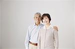 Senior adult couple cuddling and looking at camera in front of a white wall
