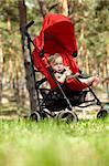 Outdoor portrait of the little boy sitting in the red buggy, eating doughnut-shaped bread roll and looking to the camera. Natural light, real colors, shallow DOF (prime 35mm L lense).