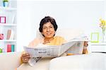 Mature 50s Indian woman reading news paper on sofa at home