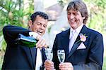 Gay couple pouring champagne to toast at their wedding.  The groom on the right is covered with champagne from the splash when the cork popped.