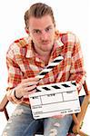 Man holding a film slate sitting down in a directors chair. White background.