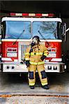 Woman firefighter stands in front of fire truck at fire station.  She is wearing bunking gear complete with gas mask and helmet.