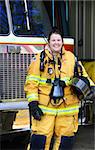 Fire Woman stands in front of fire truck wearing bunking uniform and holding helmet.  She is laughing as she is fully suited-up and ready for any fire emergency.