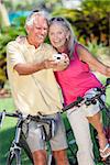 Happy senior man and woman couple together cycling on bicycles taking self portrait picture photograph with digital camera in a sunny green park