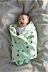 newborn baby girl in a white undershirt yawning on a bed swaddled in a baby blanket