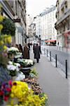 Selective focus on Man in long black coat with cane and hat walking down Paris sidewalk past colourful flower stall and other shops, along with other pedestrians