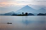 A woman paddles a dugout canoe across Lake Mutanda with its stunning backdrop of the Virunga Volcanoes, Uganda, Africa