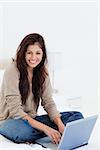 A woman sits on her bed with her laptop as she looks to the side smiling, with her legs crossed.