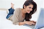 A smiling woman lying down the bed in front of her laptop with her legs raised slightly