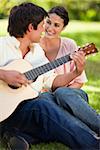 Woman smilng while looking at her friend who is playing the guitar as they both sit on the grass