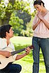 Woman standing and smiling with her hands joined as she admires her friend who is playing the guitar