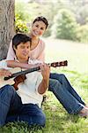 Woman sitting behind her friend and resting her arm on his chest while he plays a guitar under a tree