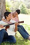 Woman looking at her friend and resting her arm on his chest while he plays a guitar under a tree