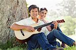 Man and his friend look straight ahead as they listen to him playing the guitar while sitting against the trunk of a tree