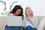 A woman holding a bank card is sitting on a couch with her friend who has a laptop