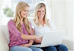 Two smiling women using a laptop as one points to the screen while they sit on the couch together