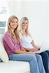 Two smiling sisters look at the camera while holding a laptop as they sit on the couch