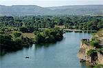 India, Rajasthan, Bhainsrorgarh. A village boat crosses a tributary of the Chambal River by the ancient crumbling walls of Bhainsrorgarh Fort.