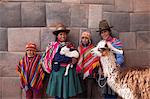 South America, Peru, Cusco. Quechua people standing in front of an Inca wall, holding a lamb and a llama and wearing traditional clothing including a bowler hat, liclla, chullo and poncho    while talking on a cell phone in the UNESCO World Heritage listed former Inca capital of Cusc