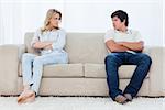 A angry couple sit at the two ends of the couch with their arms folded looking at each other