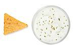 Bowl of dip with herbs beside a nacho against a white background