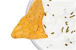 A nacho dipped into a bowl of white dip with herbs against a white background