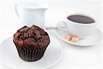 Chocolate muffin and a cup of coffee on white plates with sugar and milk against a white background