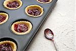 Filled, uncooked jam tarts in a tin, with a jam-covered teaspoon beside