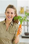 Happy young housewife holding carrot in kitchen