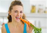 Happy young woman eating carrot in kitchen