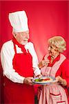 Chef teaching a sweet elderly grandma how to cook authentic Italian spaghetti marinara.  Red background with copy space.