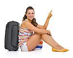 Happy young tourist woman sitting near wheel bag and pointing on copy space