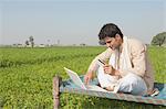 Farmer using a laptop and holding a credit card in the field, Sonipat, Haryana, India