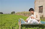 Farmer using a laptop and holding a credit card in the field, Sonipat, Haryana, India