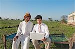 Businessman sitting in the field near his father and using a laptop, Sonipat, Haryana, India