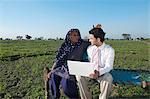 Businessman sitting in the field near his mother and using a laptop, Sonipat, Haryana, India