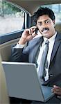 South Indian businessman using a laptop and talking on a mobile phone in the car