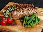 Photo of a thick sirloin steak dinner with rosemary, cherry tomatoes and green beans on a wooden board.