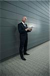 Mature businessman standing in front of wall, looking at tablet computer