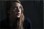 Portrait of young woman behind window, wet with raindrops, looking up