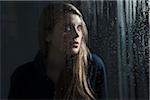 Portrait of young woman behind window, wet with raindrops, looking up