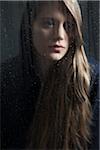 Portrait of young woman behind window, wet with raindrops, wearing hoodie