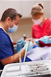 Dental cleaning tools on a white tray in the foreground. Male dentist and female dental hygienist working on patient, blurred in the background.